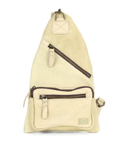 The Bed|Stü Andie is a beige leather sling bag featuring two zippered pockets and an adjustable strap, shown standing upright against a white background.