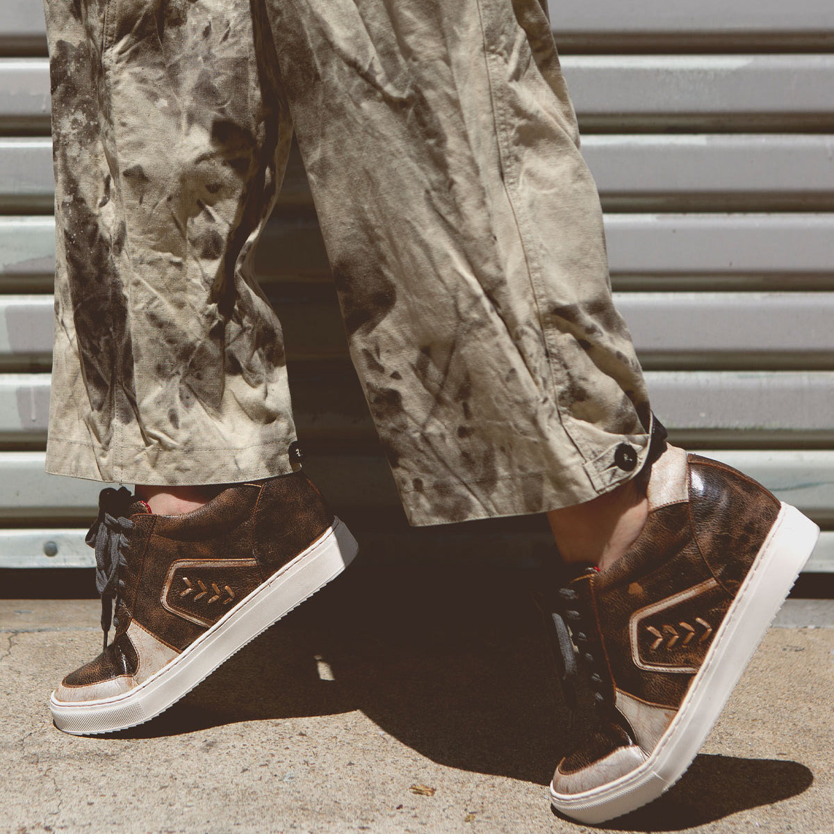 A person wearing tan, paint-splattered pants and Bed|Stü Andromeda brown high-top sneakers with white soles stands on concrete in front of a metal garage door.