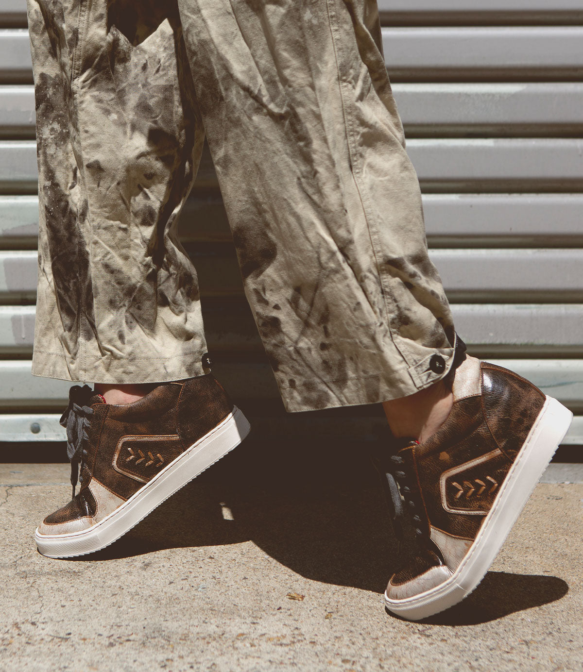 A person wearing tan, paint-splattered pants and Bed|Stü Andromeda brown high-top sneakers with white soles stands on concrete in front of a metal garage door.
