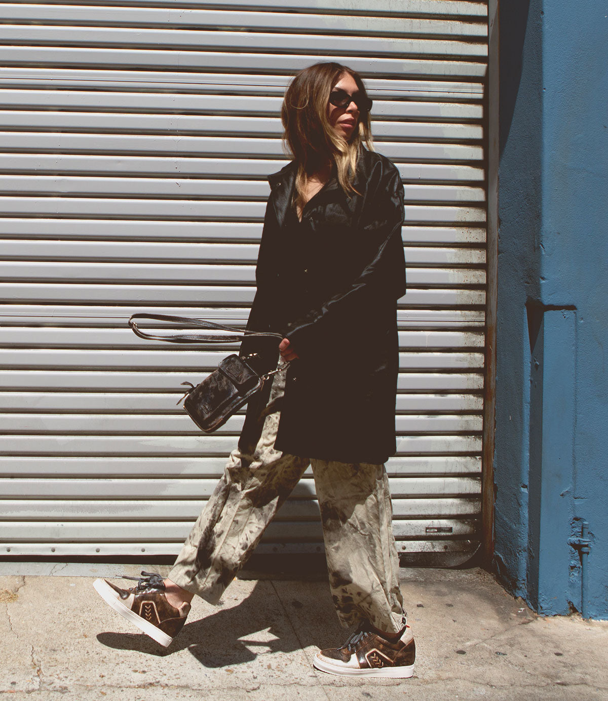 Wearing Bed|Stü’s Andromeda sneakers, a person in sunglasses, a long black coat, and patterned pants strides confidently past a corrugated metal gate, holding a small purse—city-ready style at its finest.