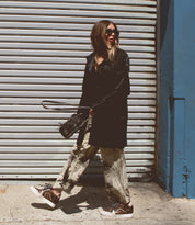 Wearing Bed|Stü’s Andromeda sneakers, a person in sunglasses, a long black coat, and patterned pants strides confidently past a corrugated metal gate, holding a small purse—city-ready style at its finest.