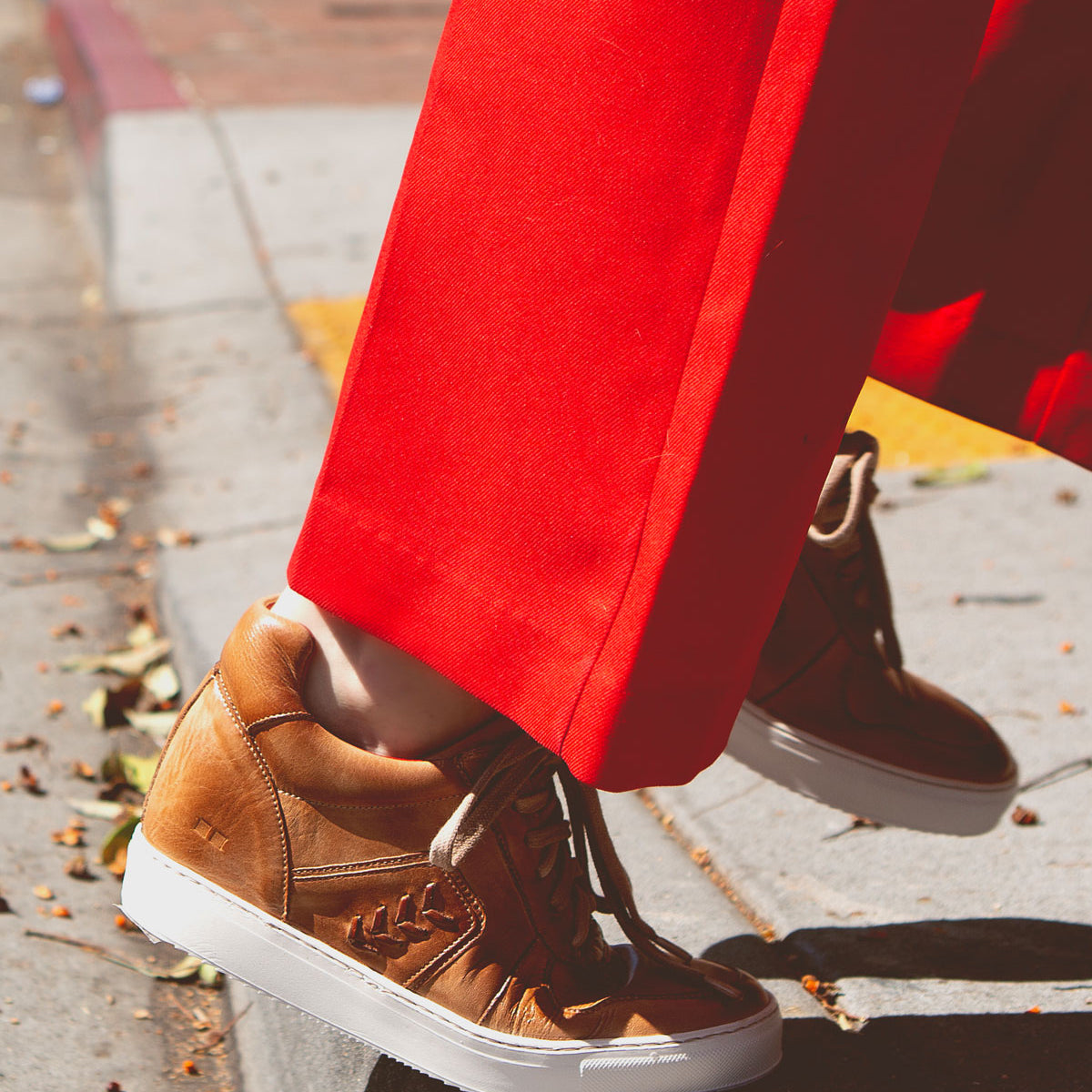 Wearing the Bed|Stü Andromeda tan city-ready sneakers, a person in bright red pants steps off a curb onto a sidewalk scattered with dry leaves.