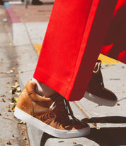 Wearing the Bed|Stü Andromeda tan city-ready sneakers, a person in bright red pants steps off a curb onto a sidewalk scattered with dry leaves.