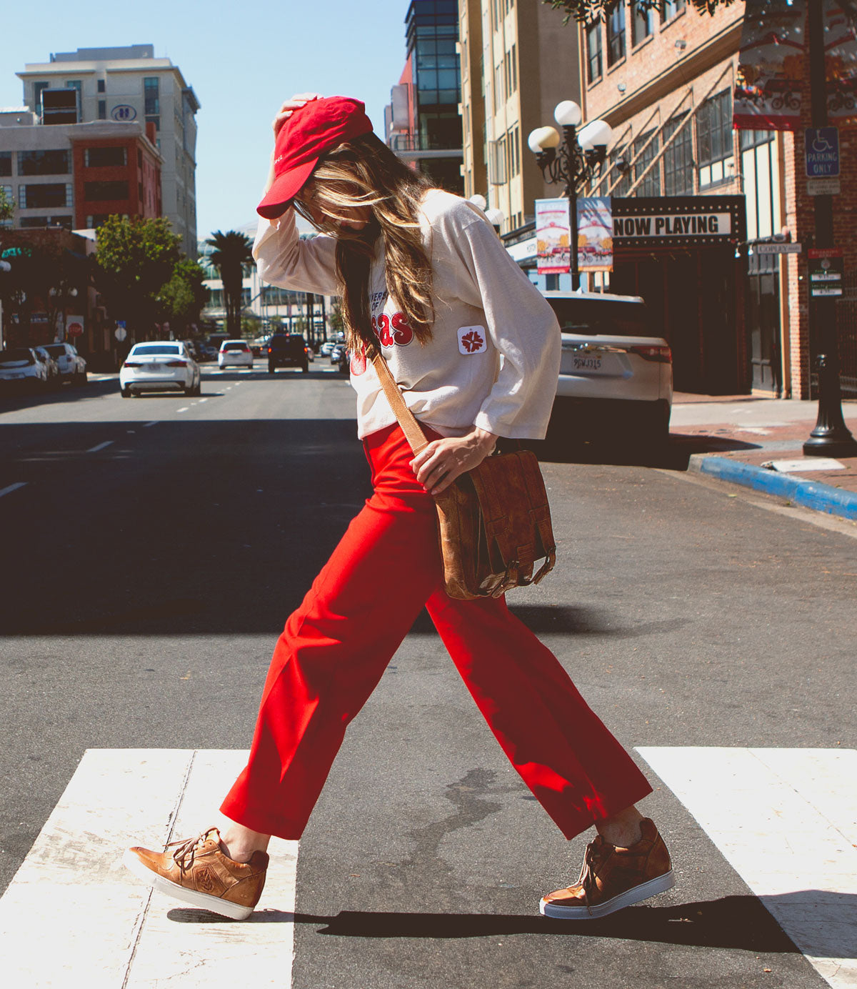 Wearing red pants, a white sweatshirt, a red cap, and brown shoes, this city stroller strides confidently across the crosswalk with a brown shoulder bag in hand—perfectly styled for urban living in Bed|Stü’s classic Andromeda sneakers.