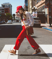 Wearing red pants, a white sweatshirt, a red cap, and brown shoes, this city stroller strides confidently across the crosswalk with a brown shoulder bag in hand—perfectly styled for urban living in Bed|Stü’s classic Andromeda sneakers.