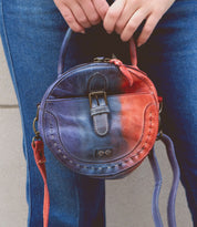 A person in blue jeans holds the Arenfield round leather crossbody bag by Bed|Stü, featuring a blue and red gradient design, a small front buckle, and an adjustable strap.