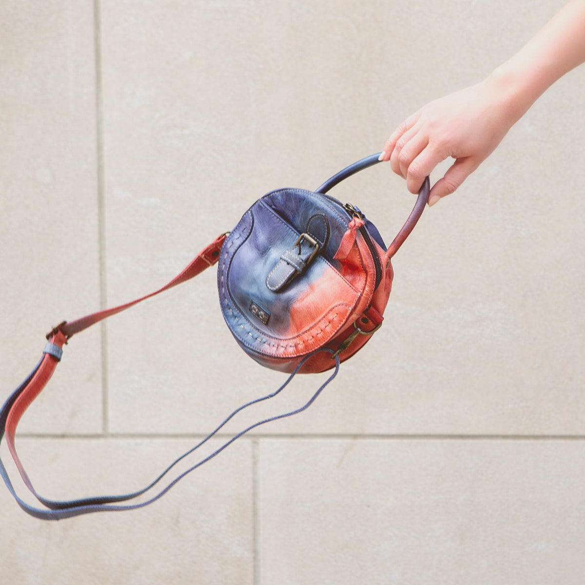 A hand holds the Bed|Stü Arenfield, a small round leather crossbody bag with a blue and red gradient design, featuring an adjustable strap and secure zip-top closure, against a neutral stone background.
