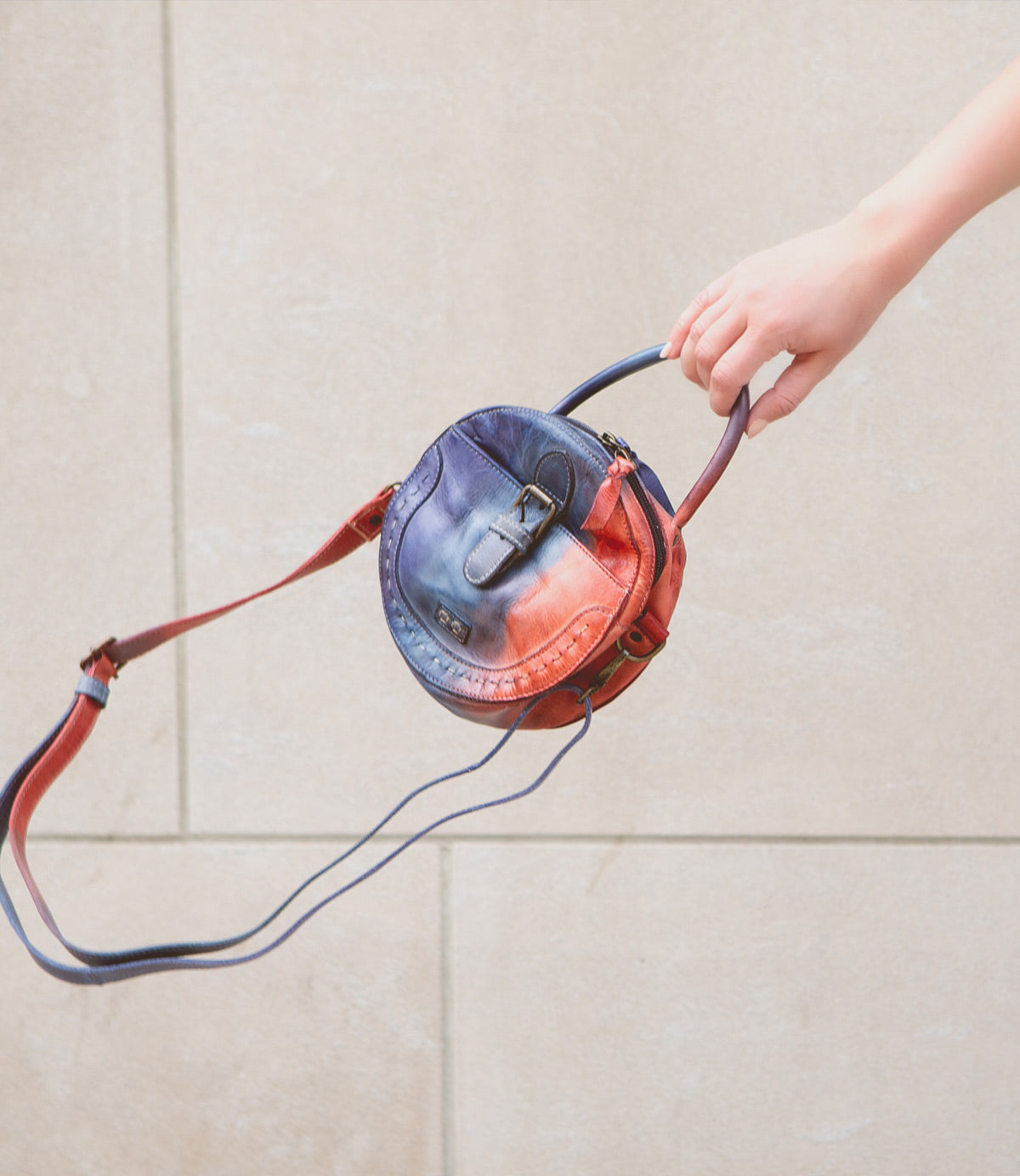 A hand holds the Bed|Stü Arenfield, a small round leather crossbody bag with a blue and red gradient design, featuring an adjustable strap and secure zip-top closure, against a neutral stone background.