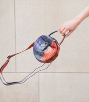 A hand holds the Bed|Stü Arenfield, a small round leather crossbody bag with a blue and red gradient design, featuring an adjustable strap and secure zip-top closure, against a neutral stone background.
