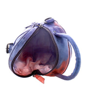 The Arenfield bag by Bed|Stü is a plush, heart-shaped accessory in purple and pink tones, featuring a round handle, adjustable strap, and zipper pull. The bag is shown partially unzipped to reveal its interior.