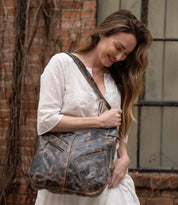 A woman in a white dress stands outdoors, smiling and looking down as she holds the Bed|Stü Argus handcrafted leather shoulder bag with oversized front pockets. A brick wall and window are visible in the background.