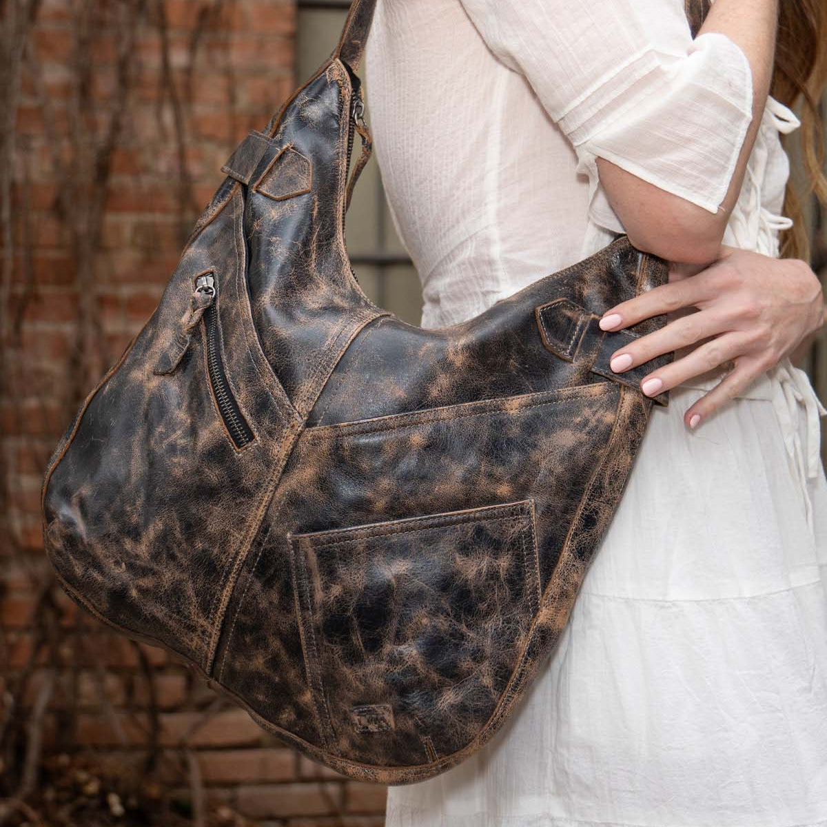 A woman in a white dress holds the Bed|Stü Argus handcrafted leather shoulder bag, featuring oversized front pockets, outdoors against a backdrop of brick wall and vines.