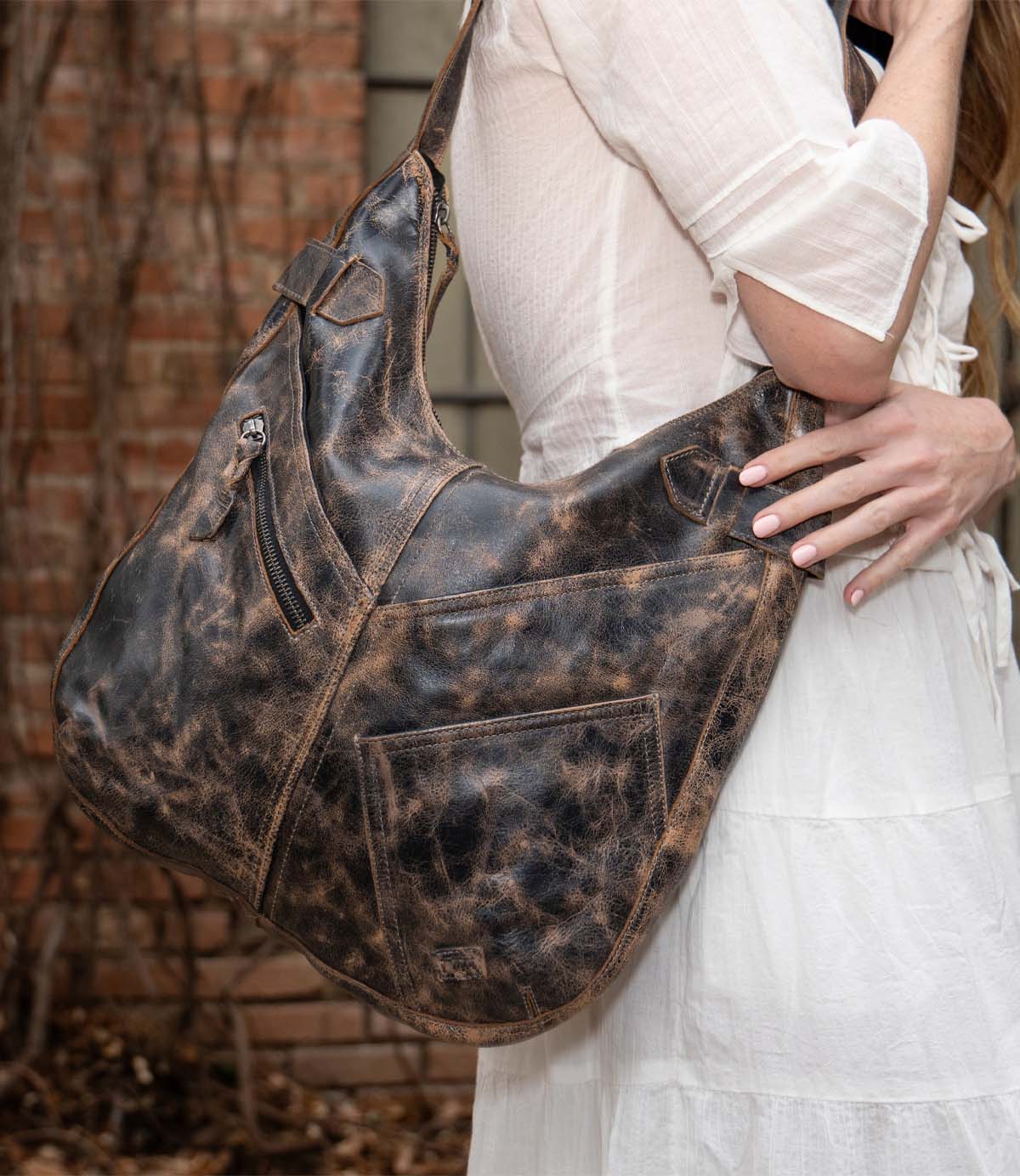 A woman in a white dress holds the Bed|Stü Argus handcrafted leather shoulder bag, featuring oversized front pockets, outdoors against a backdrop of brick wall and vines.