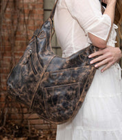 A woman in a white dress holds the Bed|Stü Argus handcrafted leather shoulder bag, featuring oversized front pockets, outdoors against a backdrop of brick wall and vines.