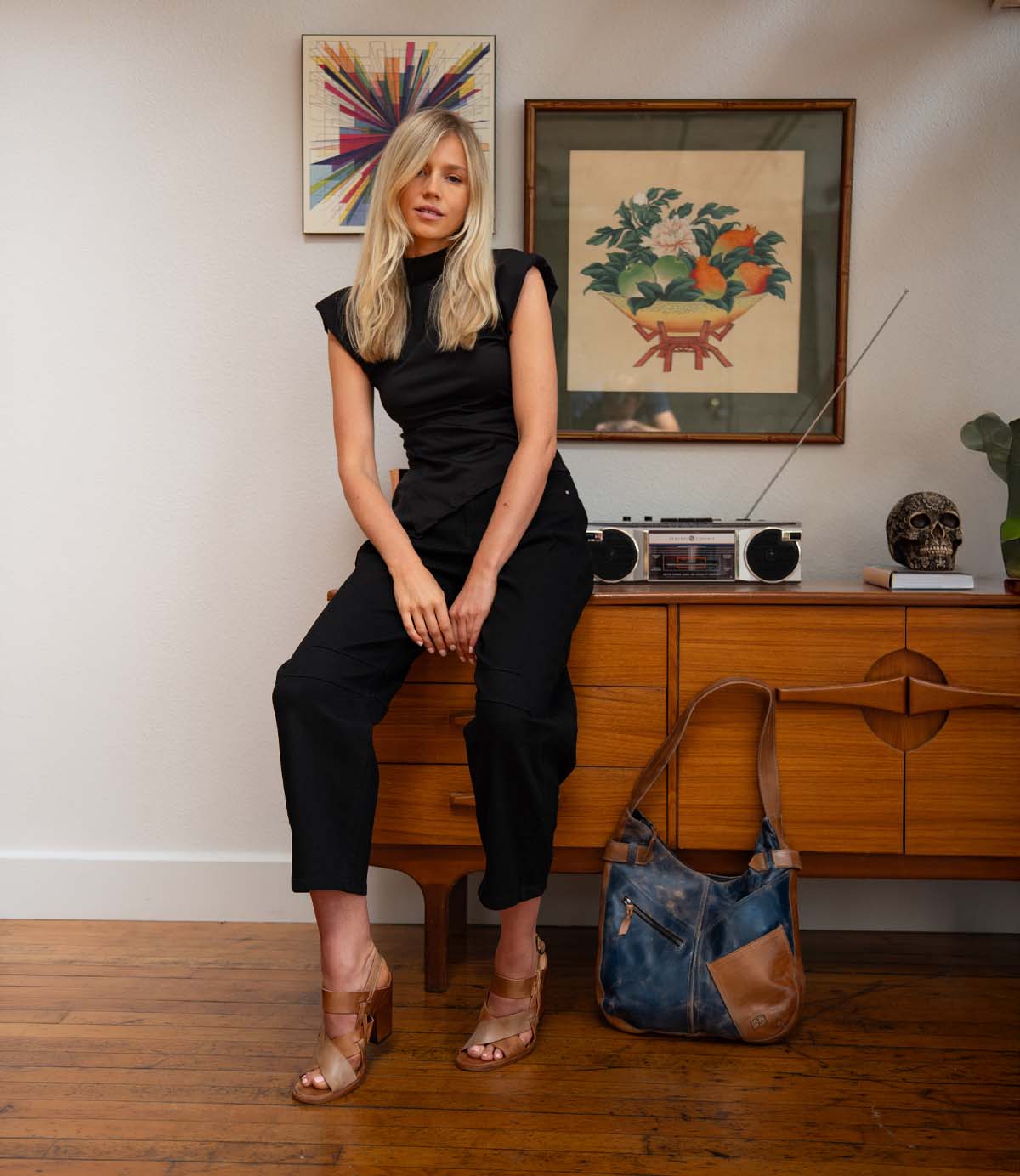 A woman dressed in black sits on a wooden console, with the Bed|Stü Argus patchwork leather bag—featuring oversized front pockets—resting on the floor beside her. Art, a radio, and decorative pieces are arranged on the wall and console behind her.