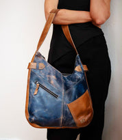 A person with arms crossed holds the Bed|Stü Argus, a large blue and brown handcrafted leather shoulder bag featuring an external zipper pocket, oversized front pockets, and a patch pocket.
