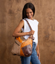 A woman stands against a brown backdrop, wearing a white sleeveless top and jeans, smiling as she looks down while holding the Bed|Stü Argus—the large handcrafted leather shoulder bag featuring oversized front pockets.