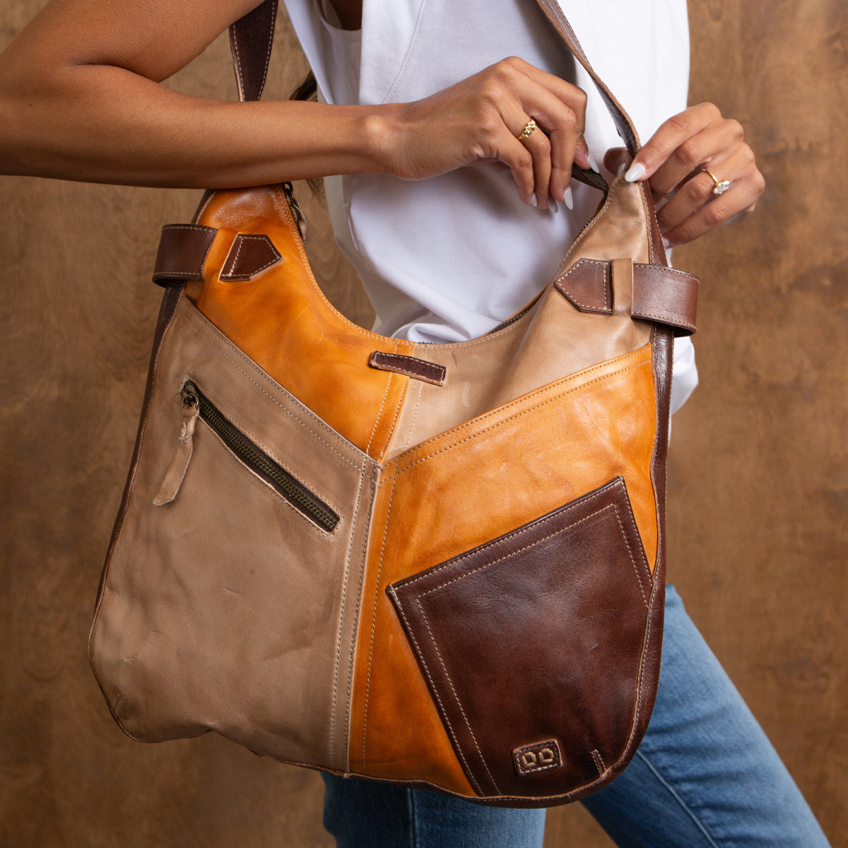 A person holds the Bed|Stü Argus, a large, handcrafted leather shoulder bag featuring a zipper and oversized front pockets, while wearing a white sleeveless top and blue jeans against a brown background.