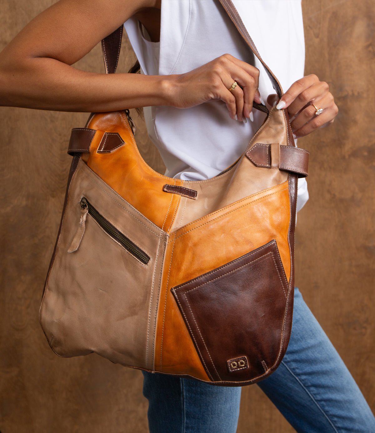A person holds the Bed|Stü Argus, a large, handcrafted leather shoulder bag featuring a zipper and oversized front pockets, while wearing a white sleeveless top and blue jeans against a brown background.