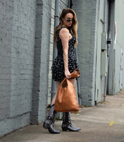 A woman in a black patterned dress, black and white boots, and sunglasses holds the Bed|Stü Ariel EM limited edition tote while standing on a city sidewalk near a gray brick building.