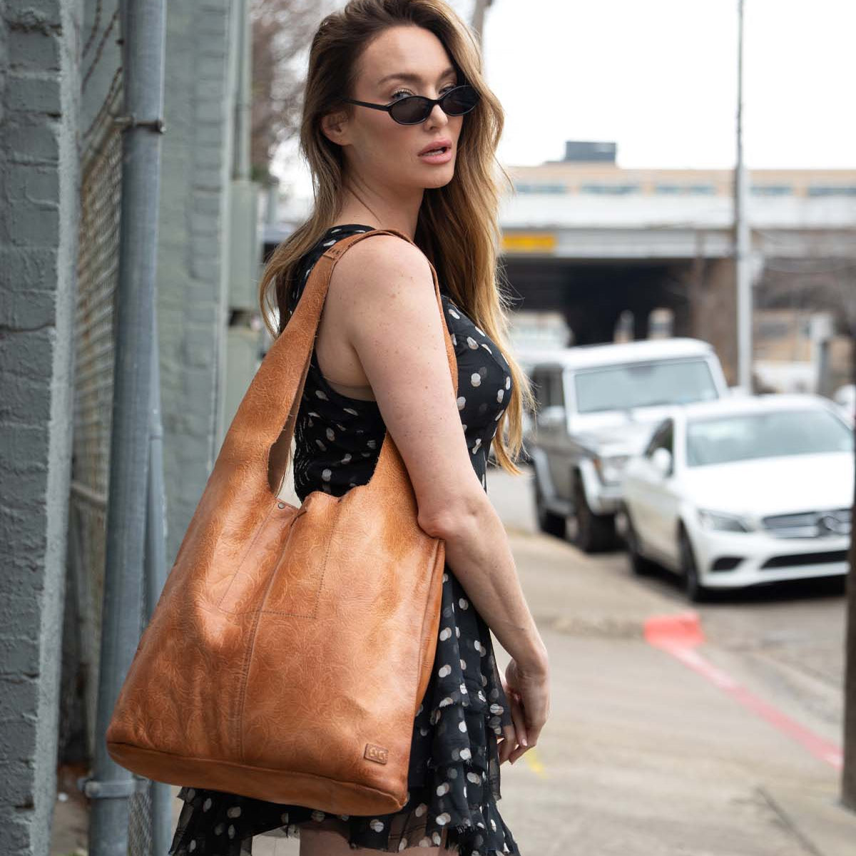 A woman in a black polka dot dress and sunglasses stands on a sidewalk, carrying the limited edition Bed|Stü Ariel EM tote over her shoulder.
