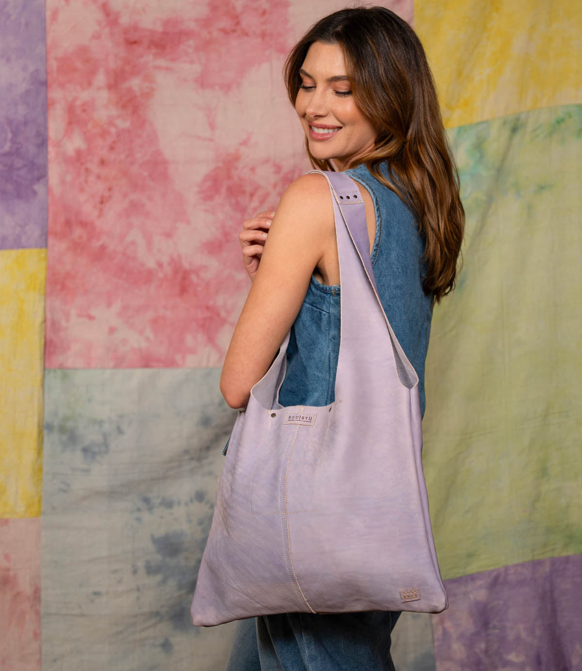 A woman in a sleeveless denim outfit smiles while holding the spacious Ariel II lavender tote bag from Bed|Stü over her shoulder. The background features pastel patchwork fabric.