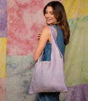 A woman in a sleeveless denim outfit smiles while holding the spacious Ariel II lavender tote bag from Bed|Stü over her shoulder. The background features pastel patchwork fabric.