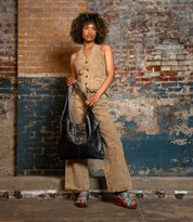 A person stands against a worn brick wall, wearing a beige vest and pants, holding a spacious leather tote, and wearing the Ariel II blue platform sandals by Bed|Stü.
