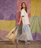 A woman in a beige t-shirt and sheer lace skirt walks, carrying the spacious Ariel II tote in matching beige from Bed|Stü. Colorful tie-dye fabric panels hang in the background.