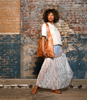 A woman stands in front of a brick wall, wearing a light-colored t-shirt, a white lace skirt, and brown sandals. She carries the Bed|Stü Ariel II, a spacious tote bag crafted from supple brown leather.