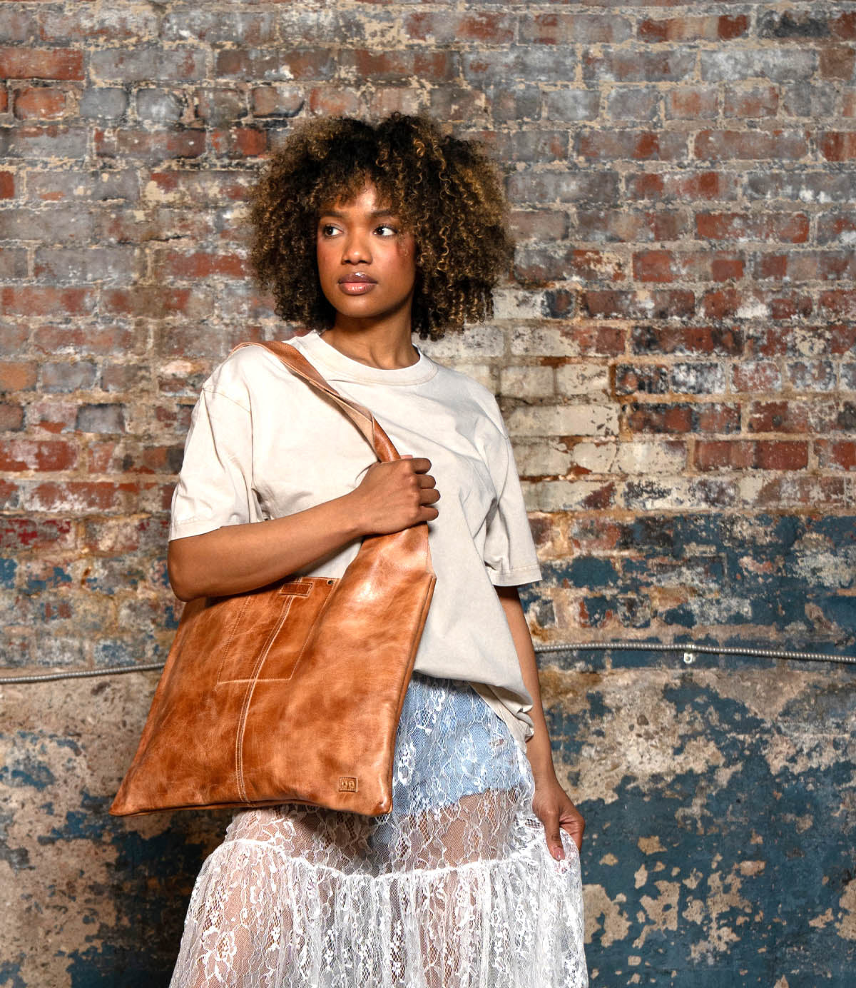 A woman stands in front of a worn brick wall, wearing a beige shirt and sheer white lace skirt, carrying the spacious Bed|Stü Ariel II leather tote bag over her shoulder.