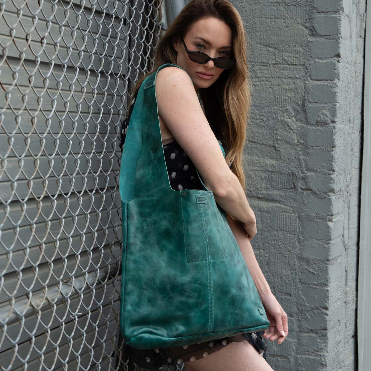 A woman in a black polka dot dress, wearing sunglasses, stands by a chain-link fence and gray brick wall, holding the Bed|Stü Ariel Max—an oversized tote with a spacious interior, crafted from hand-finished teal leather.