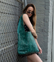 A woman in a black polka dot dress, wearing sunglasses, stands by a chain-link fence and gray brick wall, holding the Bed|Stü Ariel Max—an oversized tote with a spacious interior, crafted from hand-finished teal leather.