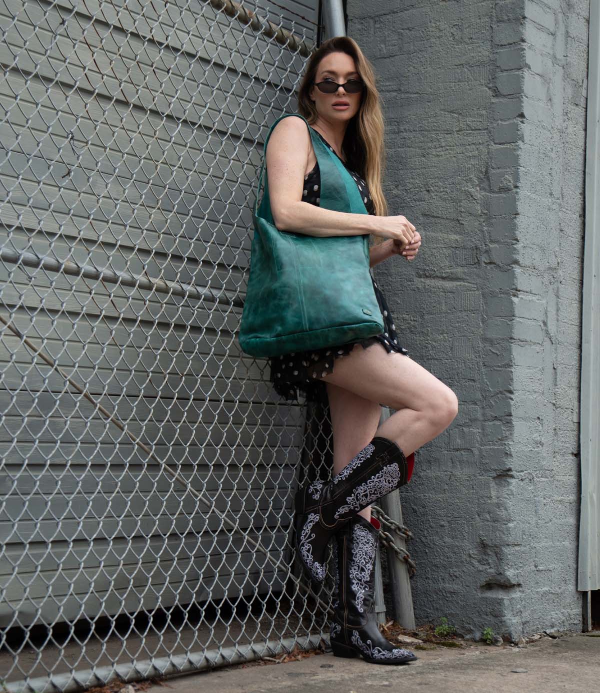 A woman in sunglasses and a black lace-trim dress stands against a chain-link fence, holding the spacious Ariel Max tote by Bed|Stü and wearing black and white cowboy boots.