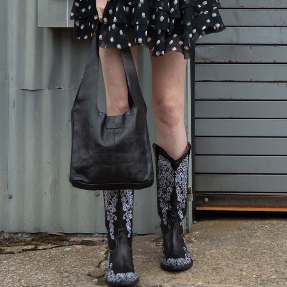 A person wearing the Ariel Mid by Bed|Stü—an artisanal shoulder bag crafted from vegetable-tanned leather—is seen standing on concrete near a metal wall and garage door, paired with a black patterned skirt and black and white embroidered cowboy boots.