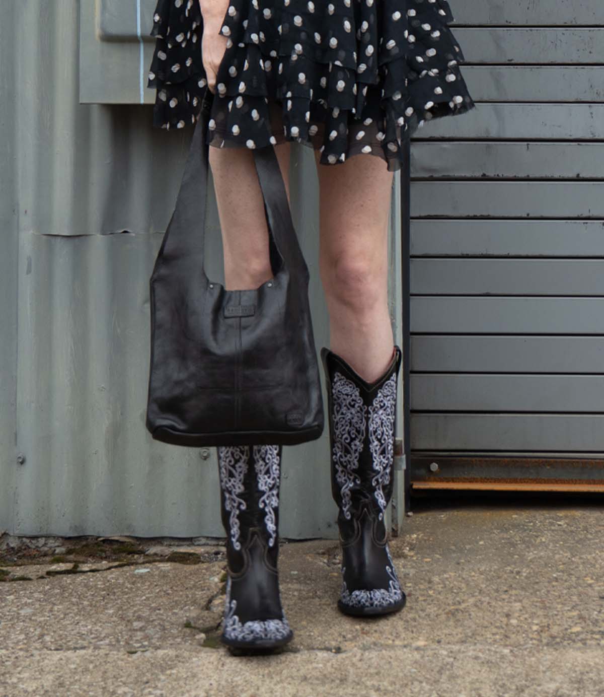 A person wearing the Ariel Mid by Bed|Stü—an artisanal shoulder bag crafted from vegetable-tanned leather—is seen standing on concrete near a metal wall and garage door, paired with a black patterned skirt and black and white embroidered cowboy boots.