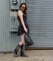 A woman in a black polka dot dress, black patterned cowboy boots, and sunglasses stands holding the Ariel Mid artisanal shoulder bag by Bed|Stü, crafted from vegetable-tanned leather, in front of a gray industrial garage door.