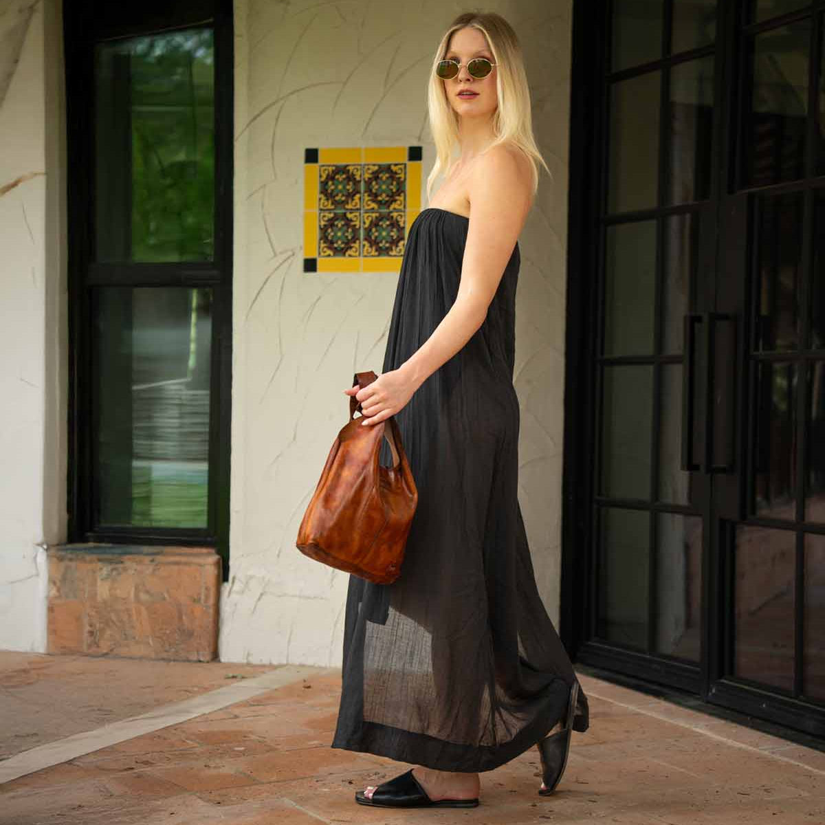 A woman in a strapless black dress, black sandals, and sunglasses stands outdoors near a wall and glass doors, holding the Bed|Stü Ariel Mid artisanal shoulder bag.