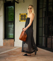 A woman in a strapless black dress, black sandals, and sunglasses stands outdoors near a wall and glass doors, holding the Bed|Stü Ariel Mid artisanal shoulder bag.