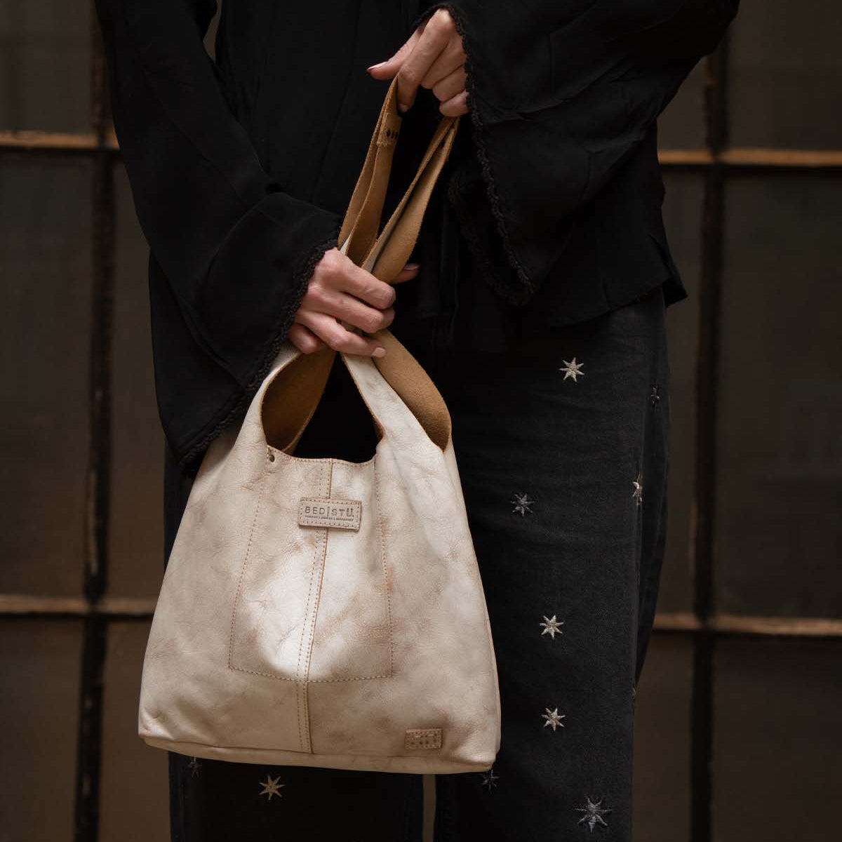 A person wearing a black top and star-patterned black pants holds the Bed|Stü Ariel Mid, an artisanal shoulder bag crafted from beige vegetable-tanned leather, in front of a window with wooden frames.