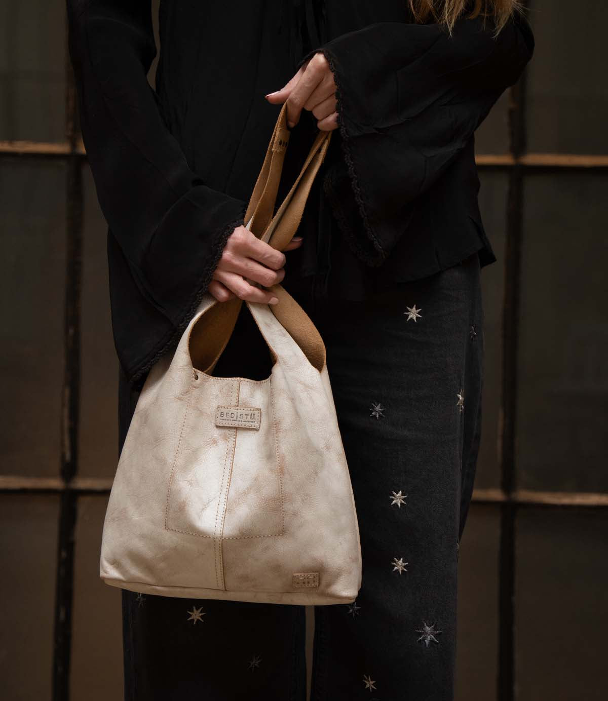 A person wearing a black top and star-patterned black pants holds the Bed|Stü Ariel Mid, an artisanal shoulder bag crafted from beige vegetable-tanned leather, in front of a window with wooden frames.