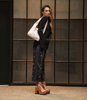 A woman stands sideways in front of a window, dressed in a black shirt, star-patterned black pants, and brown platform shoes, carrying the Bed|Stü Ariel Mid — a large artisanal shoulder bag crafted from vegetable-tanned leather — over her shoulder.