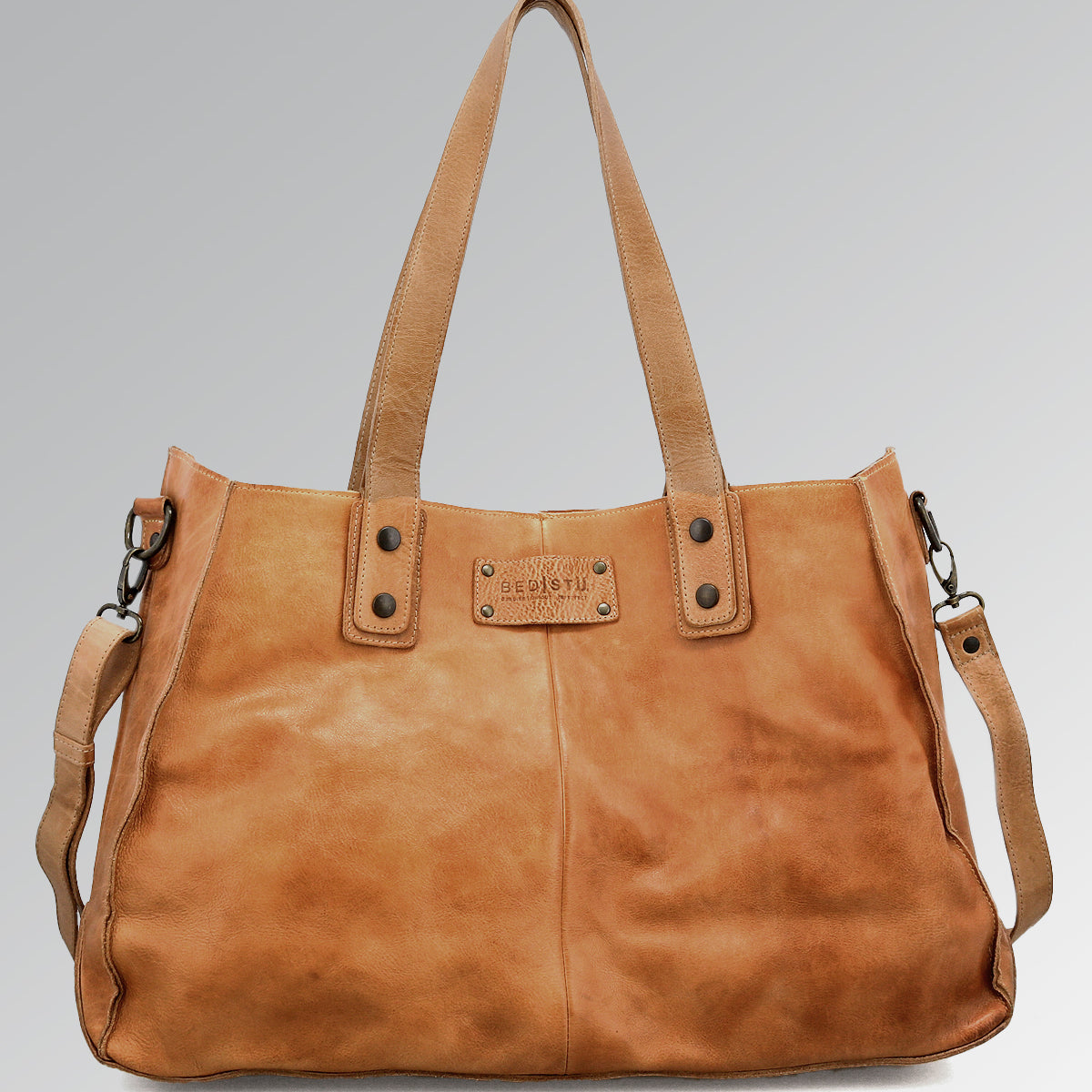 Introducing the Bed|Stü Arlington II—a spacious tan leather handbag featuring double handles, a detachable shoulder strap, and a signature rectangular metal logo plate on the front. Set against a plain gradient background, this distinctive accessory exemplifies custom craftsmanship.