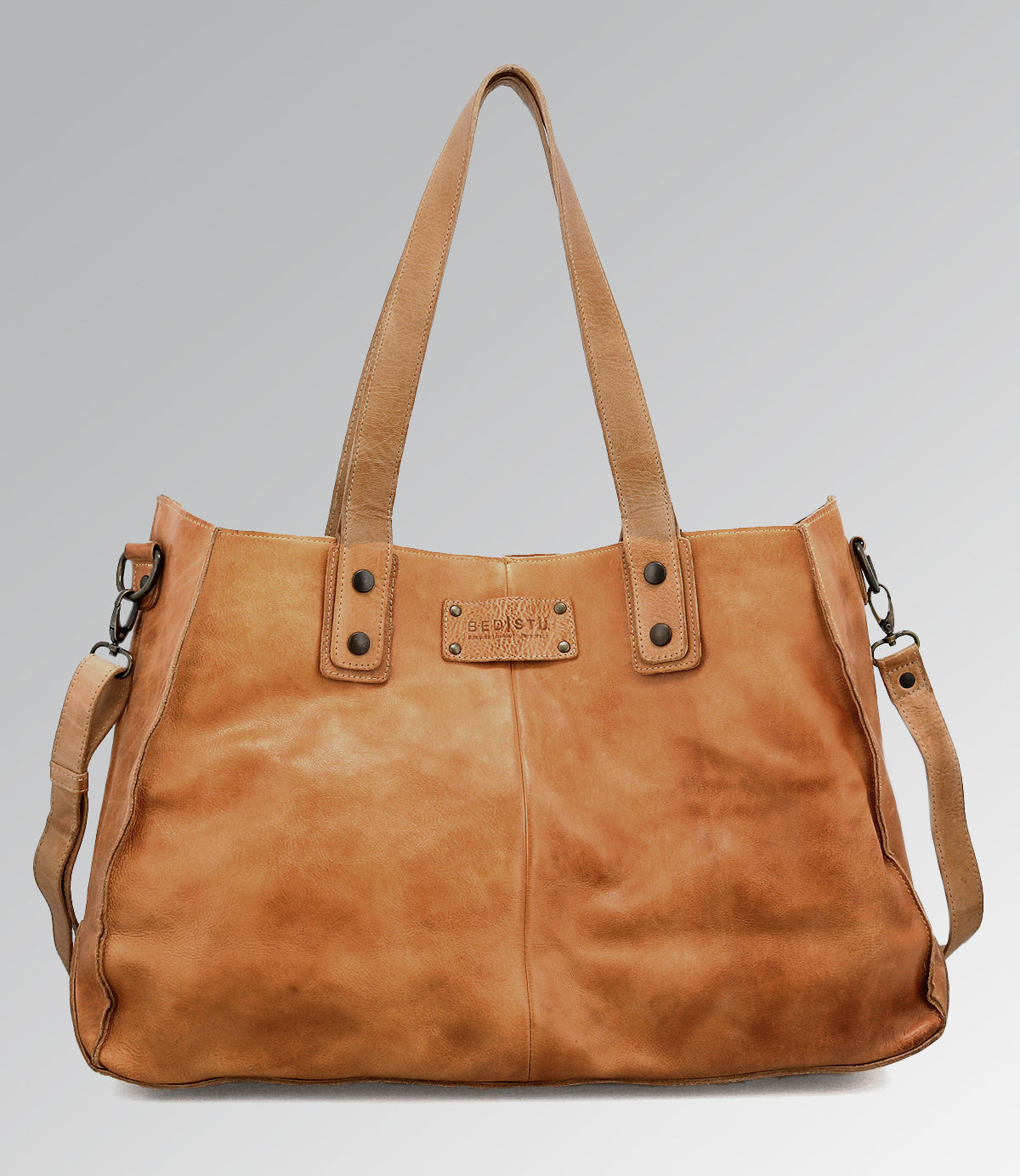 Introducing the Bed|Stü Arlington II—a spacious tan leather handbag featuring double handles, a detachable shoulder strap, and a signature rectangular metal logo plate on the front. Set against a plain gradient background, this distinctive accessory exemplifies custom craftsmanship.