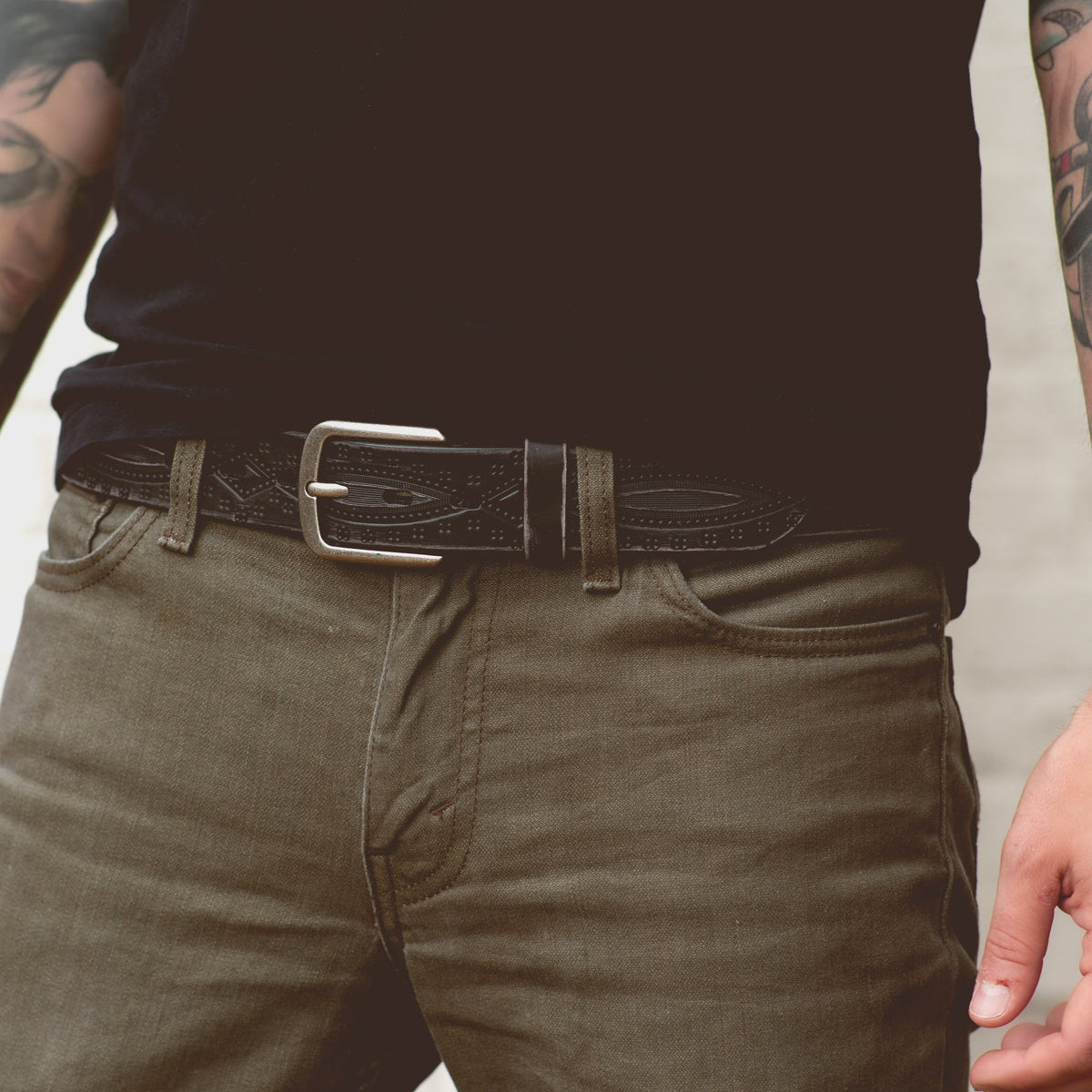A person wearing olive green jeans, a black shirt, and the Bed|Stü Arsenal genuine leather belt with a removable silver buckle. The versatile western-style belt adds character to the look. Visible forearm tattoos appear on both arms, with only the torso and upper legs shown.