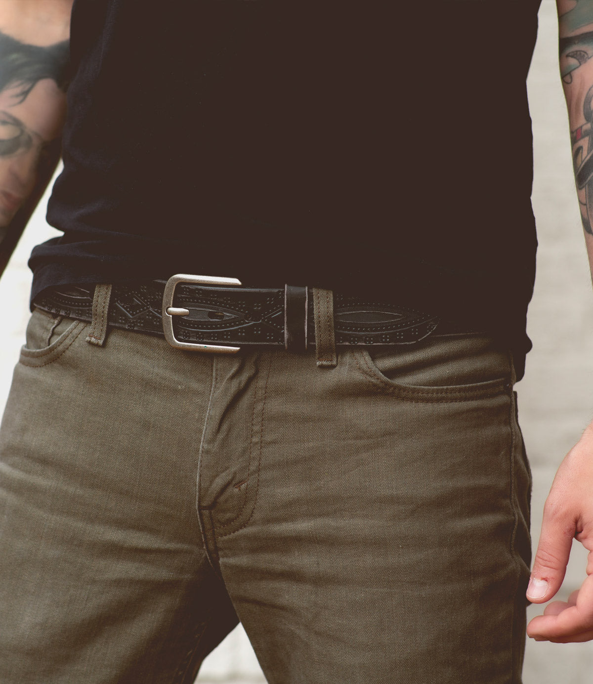 A person wearing olive green jeans, a black shirt, and the Bed|Stü Arsenal genuine leather belt with a removable silver buckle. The versatile western-style belt adds character to the look. Visible forearm tattoos appear on both arms, with only the torso and upper legs shown.