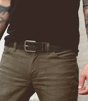 A person wearing olive green jeans, a black shirt, and the Bed|Stü Arsenal genuine leather belt with a removable silver buckle. The versatile western-style belt adds character to the look. Visible forearm tattoos appear on both arms, with only the torso and upper legs shown.