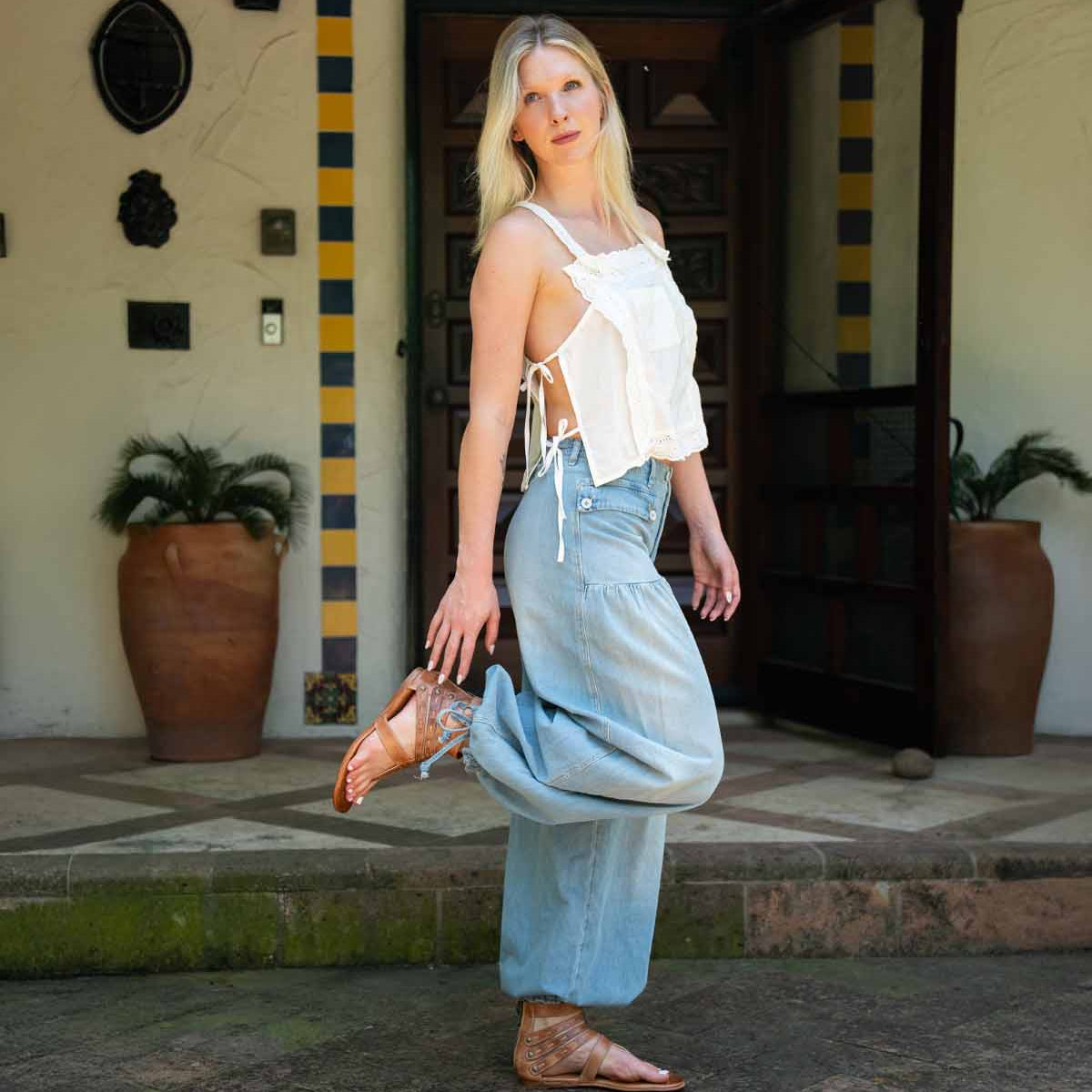 A woman stands outdoors on one leg in front of a building with potted plants and a tiled entryway, wearing a white sleeveless top, light blue wide-leg jeans, and the Bed|Stü Artemis II brown thong sandals featuring delicate leather straps.