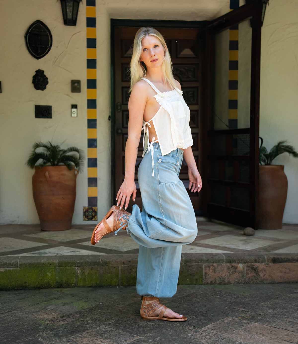 A woman stands outdoors on one leg in front of a building with potted plants and a tiled entryway, wearing a white sleeveless top, light blue wide-leg jeans, and the Bed|Stü Artemis II brown thong sandals featuring delicate leather straps.