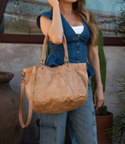 A woman wearing a blue vest and jeans holds the Bed|Stü Astana, a large tan leather everyday bag with shoulder straps, while standing in front of a window and a cactus.